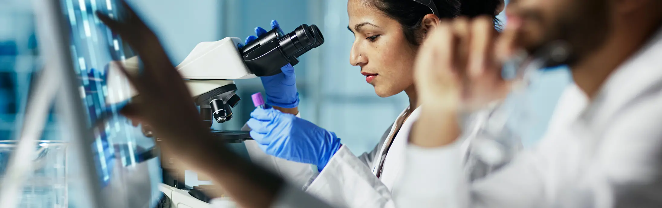 实验室里，一名女性坐在显微镜前观察样本，画面前方是一名留着胡子的男性正在看电脑屏幕