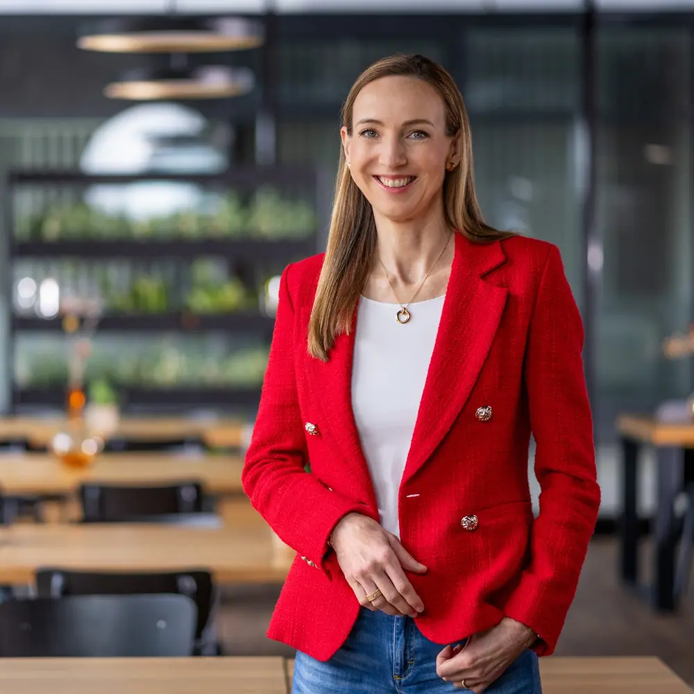 Simone Bagel-Trah博士身着亮红色西装外套和白色上衣，站在一个现代风格的咖啡馆中，背景是木质桌子和装饰花瓶。