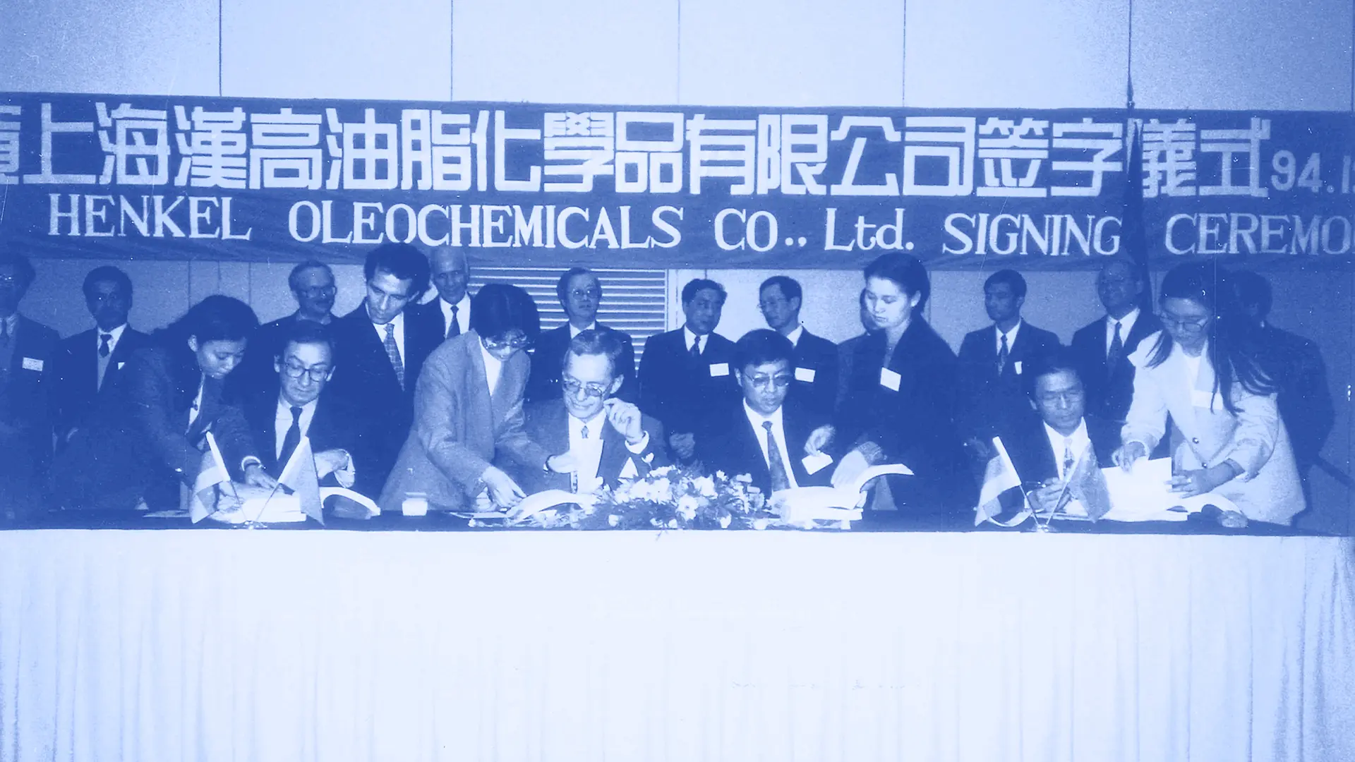 4 men in suits sitting behind the table to sign on documents with people in suits standing behind them. Image in black and white.