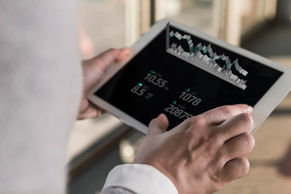 在明亮的室内环境中，一个人手持平板电脑，屏幕上展示着数字化图表和数据，图片从肩后视角拍摄。